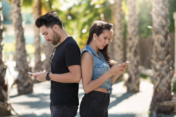 Couple standing back to back outside using mobiles