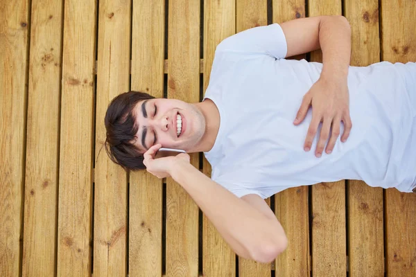 Молодой счастливый человек лежит на деревянных досках и разговаривает по мобильному телефону — стоковое фото