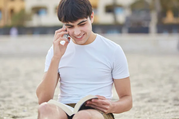 Joven hombre feliz hablando en el móvil y leyendo libro fuera — Foto de Stock
