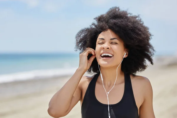 Jovem afro-americana ouvindo audiolivro na praia rindo — Fotografia de Stock