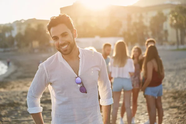 Guapo joven de pie en la playa con amigos sonriendo — Foto de Stock