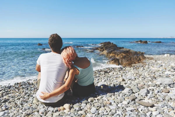 Et par som sitter på en steinete strand og ser på havet – stockfoto