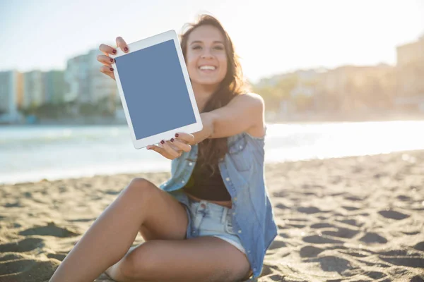 Joven mujer feliz sentado en la playa mostrando tableta — Foto de Stock