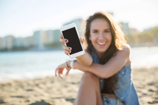 Joven mujer feliz sentada en la playa mostrando teléfono inteligente — Foto de Stock