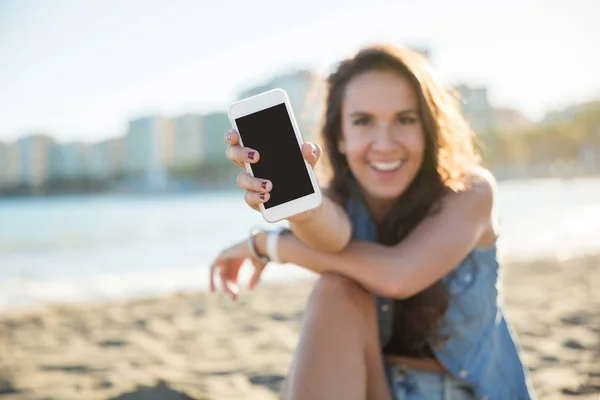 Jovem mulher feliz sentada na praia mostrando smartphone Fotos De Bancos De Imagens