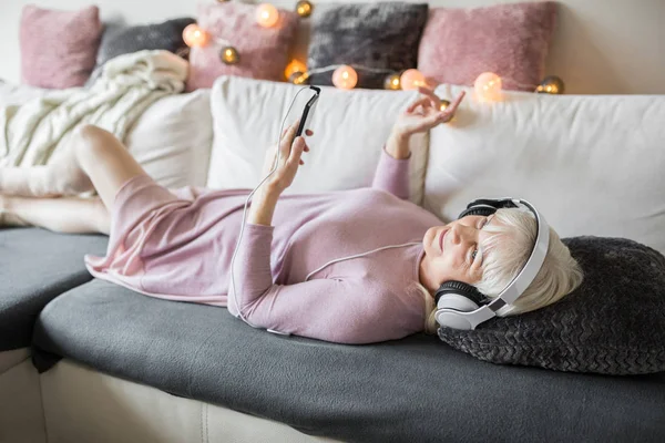 Mujer mayor disfrutando de la música en el móvil tumbado en el sofá — Foto de Stock