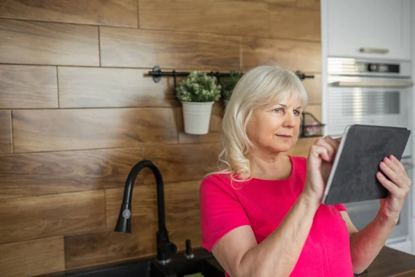 Senior senhora em vestido vermelho usando tablet na cozinha — Fotografia de Stock