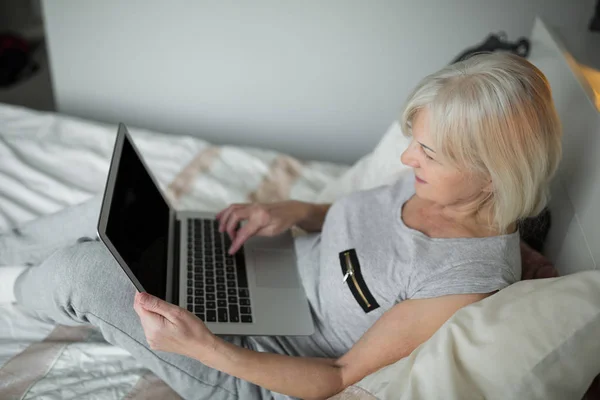 Señora mayor sentada en la cama usando portátil — Foto de Stock