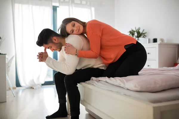 Hombre molesto sentado en la cama y su novia abrazándolo — Foto de Stock
