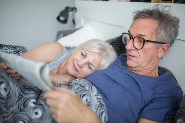 Hombre mayor acostado en la cama con su esposa leyendo el periódico — Foto de Stock