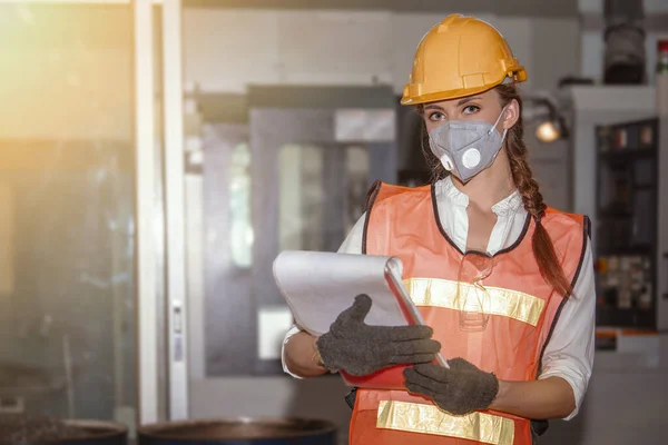 Young woman engineer in the mask protected herself with air pollution & virus  holding clipboard with blurred controller machine in factory background