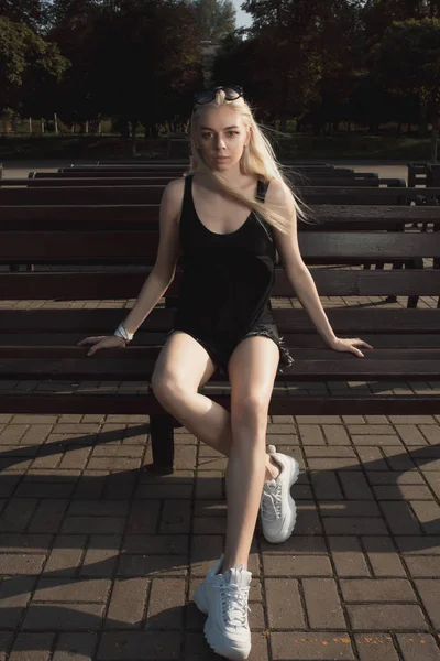 Pretty blonde model in stylish apparel posing on a bench in sunn