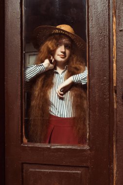 Genç ve güzel kırmızı saçlı model ahşap kapının arkasında hasır şapkalı