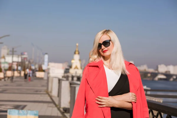 Stylish blonde model wears sunglasses, posing in red cloak on a — Stock Photo, Image