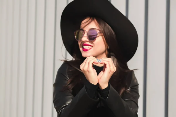 Молода здорова модель в чорному капелюсі та окулярах, що показують форму серця — стокове фото
