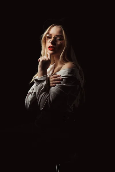 stock image Glamor blonde girl in glasses and hat, wearing shirt with naked shoulders, posing with dramatic light at studio