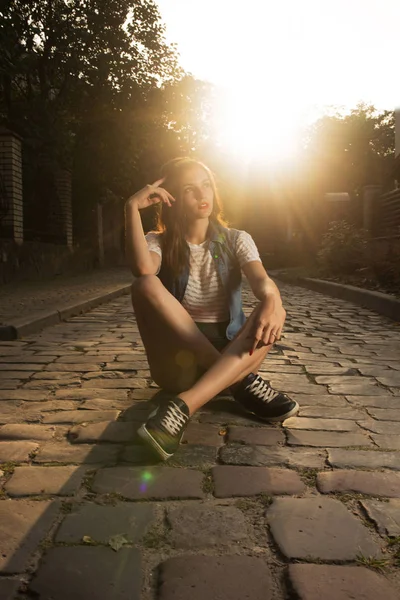 Fashionable brunette girl in sport apparel sitting on the cobbles at the evening