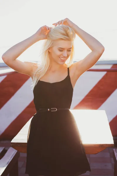 Cheerful blonde woman wears elegant black dress posing at the roof in sunny evening