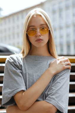 Sarışın kızın yakın plan portresi. Sarı gözlüklü ve gri tişörtlü genç kız şehirde yürüyor.