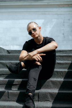 Örgülü şık Avrupalı adam sokak merdivenlerinde otururken güneş gözlüğü takıyor.
