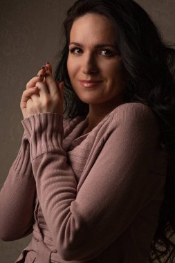 Uzun kıvırcık saçlı ve pembe makyajlı neşeli genç bir kadın gölgede örülmüş bir ceket giyiyor.