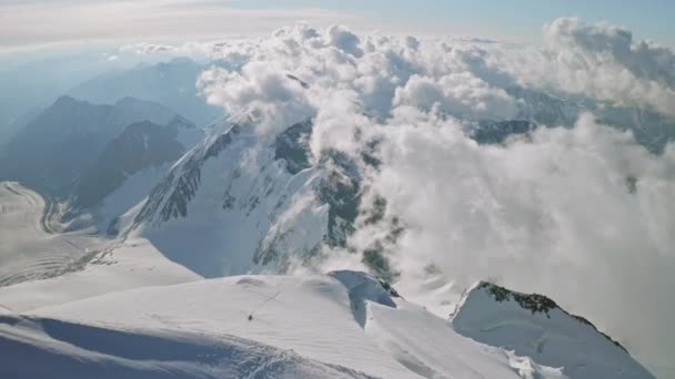 Episka magiska moln flyter under snötoppen, klättrare stiger upp till bergstoppen — Stockvideo