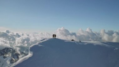 Karlı dağlarda dağcılar bulutların tepesinde, drone daireler çiziyor.