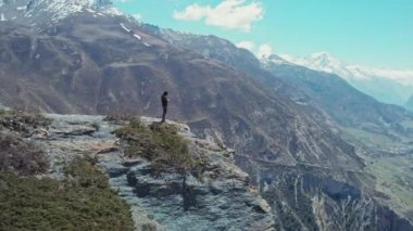 Uçurumun kenarındaki yüksek kayalıklarda duran adam, etrafındaki muhteşem güneşli dağ manzarası.