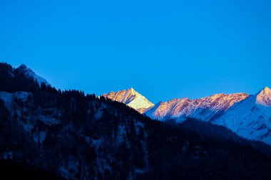 Karla kaplı bir dağın tepesinde gün doğumu. Öndeki dağ çam ağaçlarıyla kaplı..