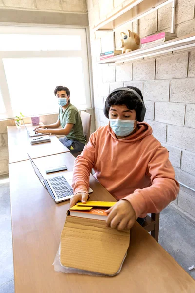 Young man with face masks receiving a package delivery of purchase made online, while doing home office, during COVID-19 pandemic.
