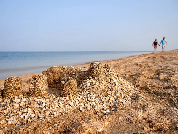Sand castle with shells on the beach against the background of two people holding hands