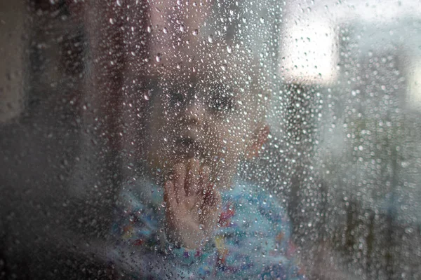 White baby boy looking through glass window on the rain drops and rain outside and pressing his small palm against the glass - isolation, quarantine, virus outside, stay safe, stay home closer portrait looking up surprised