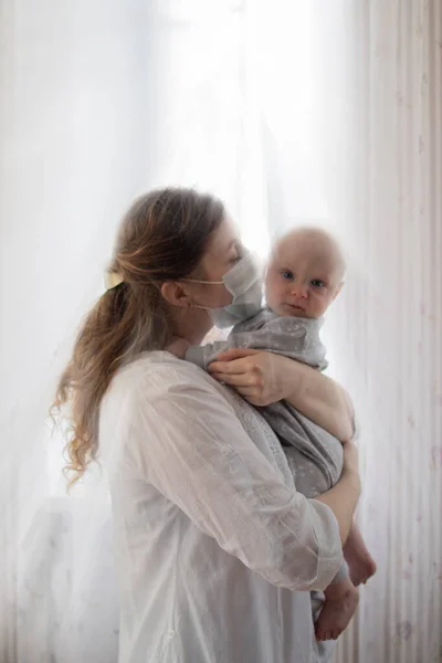 Mother or doctor wearing a face mask holding a baby boy staying inside safe in times of coronavirus pandemic spread in the world near a window surrounded by light sweetly kissing through the mask