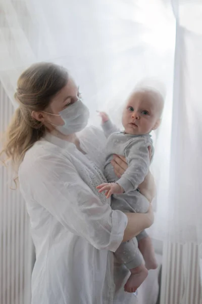 Anne ya da doktor yüz maskesi takıyor. Bebek koronavirüs salgınının olduğu zamanlarda bebeğini kucağında tutuyor. Etrafı güneş ışınlarıyla çevrili bir pencerenin yakınına yayılmış.