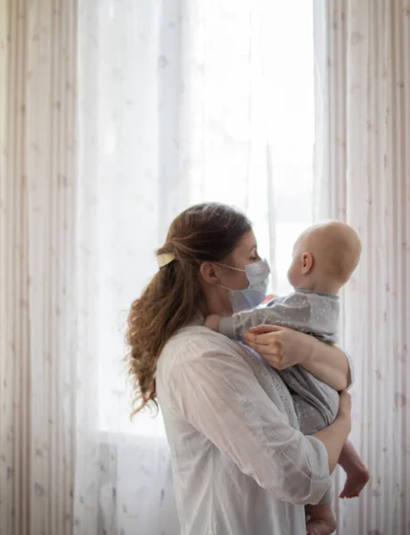 Mother or doctor wearing a face mask holding a baby boy staying inside safe in times of coronavirus pandemic spread in the world near a window surrounded by light looking at each other