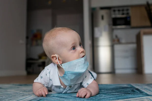 Beyaz erkek bebek İskandinav tarzı bir dairede mavi halıda yatıyor mavi bir tıbbi maskeyle oynuyor ve izolasyon anında her yöne doğru uzanıyor ve Coronavirus covid yüzünden evde kalıyor.