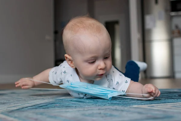 Beyaz erkek bebek İskandinav tarzı bir dairede mavi halıda yatıyor mavi bir tıbbi maskeyle oynuyor ve izolasyon anında her yöne doğru uzanıyor ve Coronavirus covid-19 yüzünden evde kalıyor.