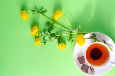 Bir fincan siyah çay, bir parça çikolata ve yeşil arka planda Trollius europaeus 'un sarı çiçekleri. Çikolata günü ve sabah çayı.