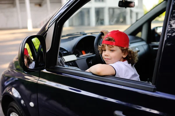 Niño con el pelo rizado y una gorra roja se sienta detrás del volante del coche — Foto de Stock