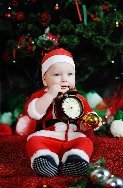 Child dressed as Santa Claus gnawing teeth clock alarm . Ch — Stock Photo, Image