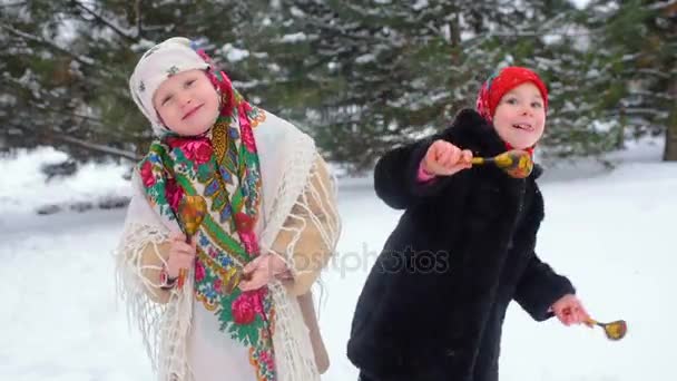 Две маленькие девочки в одежде и платках в русском стиле играют на деревянных ложках на фоне снега — стоковое видео