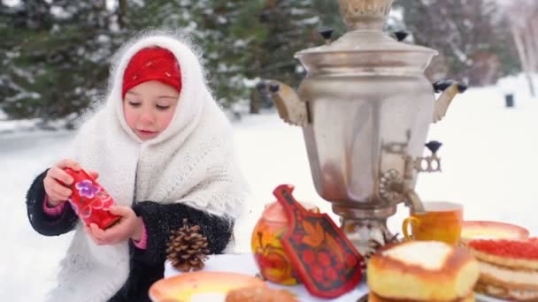 Ein kleines Mädchen in Pelzmantel und Schal im russischen Stil sitzt an einem festlichen Tisch und spielt eine russische Matrjoschka — Stockvideo