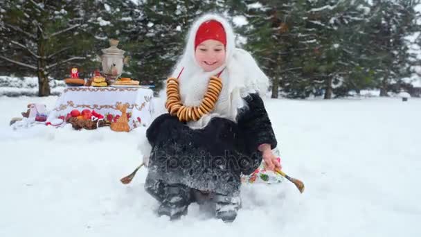 A little girl in a fur coat and a scarf in Russian style with a bunch of bagels around her neck is showered with snow — Stock Video