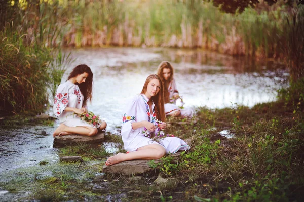 Three young beautiful girls in white shirts with floral ornaments with wreaths in their hands sitting on the background of the river. — Stock Photo, Image