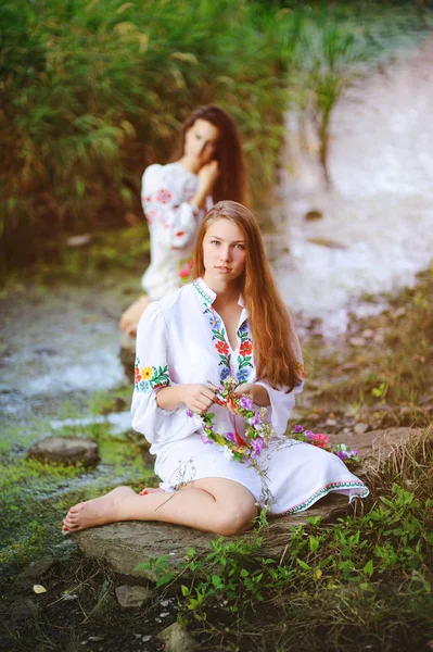Δύο νεαρά κορίτσια όμορφα σε λευκό πουκάμισο με φλοράλ στολίδι με στεφάνια στα χέρια τους, κάθεται στο παρασκήνιο του ποταμού. — Φωτογραφία Αρχείου