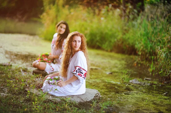 Iki genç güzel kızla çiçek süsleme ile Beyaz gömlekli çelenk nehrin arka plan üzerinde oturan ellerinde. — Stok fotoğraf