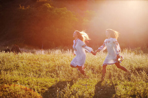 two young beautiful girls in white shirts with floral ornament with flower wreaths in their hands run against the background of nature and grass in the contour or the back light of the sun.