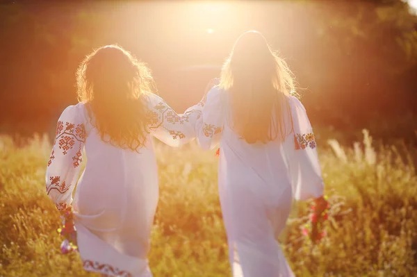 Duas meninas bonitas jovens em camisas brancas com ornamento floral com coroas de flores em suas mãos correr contra o fundo da natureza e grama no contorno ou a luz traseira do sol . — Fotografia de Stock