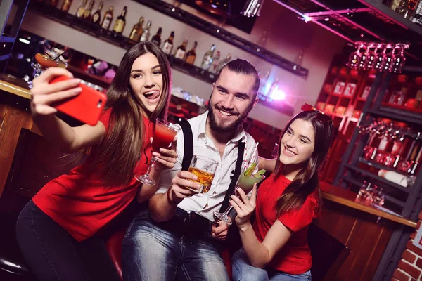 friends - two young attractive girls and a guy at a nightclub party make a photo of themselves on a mobile phone or smartphone