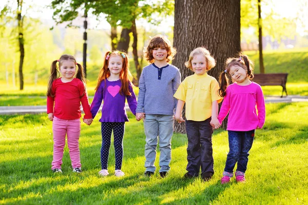 Un grupo de niños pequeños sonriendo tomados de la mano sobre un fondo de hierba, un árbol y un parque . — Foto de Stock